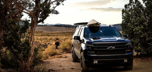 Dark Chevrolet Silverado pickup with a camper shell and roof-mounted kayak parked on a dirt road between trees.